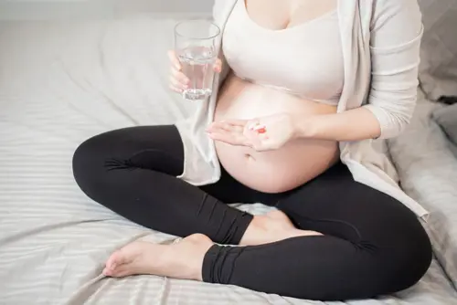Nėščia moteris sėdėdama ant lovos geria kasdienę vitaminų dozę.